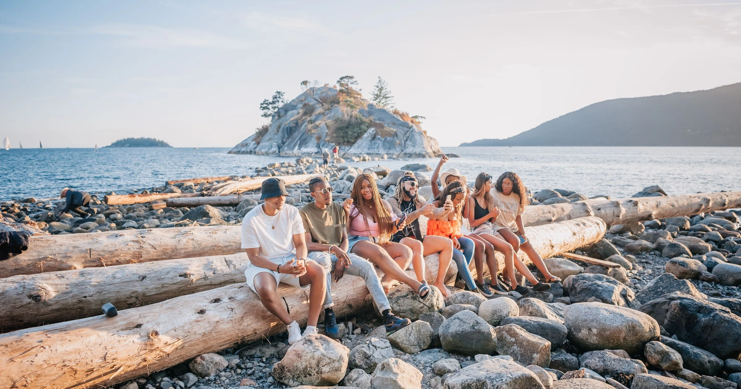 Friends relax on logs by the rocky shore, enjoying a sunny day at the beach.
